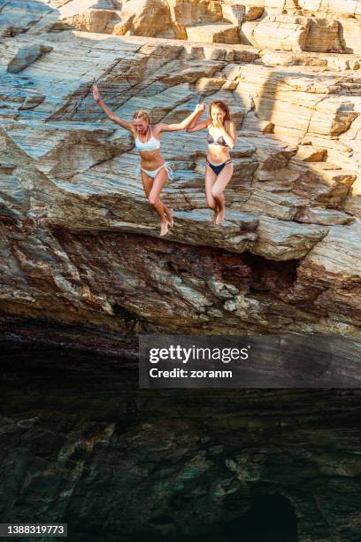 two young women in bikinis diving into sea water from cliff - cliff dive stock pictures, royalty-free photos & images