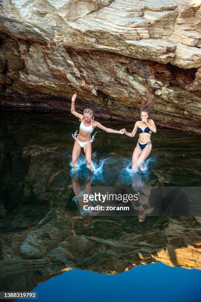 zwei junge frauen in bikinis, die sich an den händen halten und mit spritzer ins meerwasser tauchen - klippenspringen stock-fotos und bilder