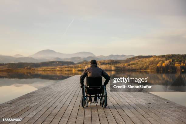 hombre discapacitado sentado en silla de ruedas en un embarcadero y admirando la naturaleza junto al lago - wheelchair fotografías e imágenes de stock