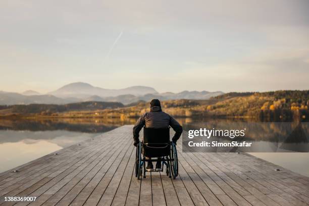 disabled man sitting in wheelchair on jetty and admiring the nature by lake - rolstoel stockfoto's en -beelden