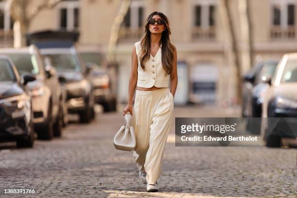 Angela Gonzalez wears black sunglasses, silver earrings, a white latte buttoned / sleeveless / linen gilet, matching white latte palazzo pants, a...