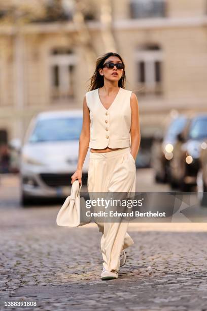 Angela Gonzalez wears black sunglasses, silver earrings, a white latte buttoned / sleeveless / linen gilet, matching white latte palazzo pants, a...