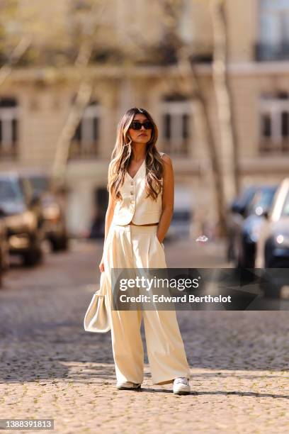 Angela Gonzalez wears black sunglasses, silver earrings, a white latte buttoned / sleeveless / linen gilet, matching white latte palazzo pants, a...