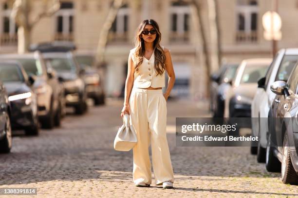 Angela Gonzalez wears black sunglasses, silver earrings, a white latte buttoned / sleeveless / linen gilet, matching white latte palazzo pants, a...