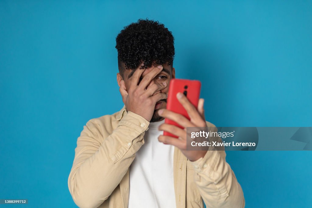 Nervous and scared latino american young man covering his face while reading bad news on the mobile phone against blue background