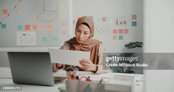 junge asiatische muslimische geschäftsfrau im hijab, die an einem laptop am schreibtisch im büro arbeitet. - economist stock-fotos und bilder