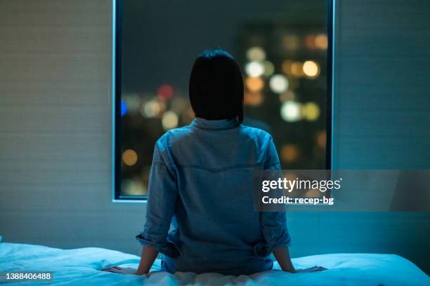 vue arrière d’une femme assise seule sur son lit dans la chambre et regardant par la fenêtre la nuit - femme fenêtre photos et images de collection