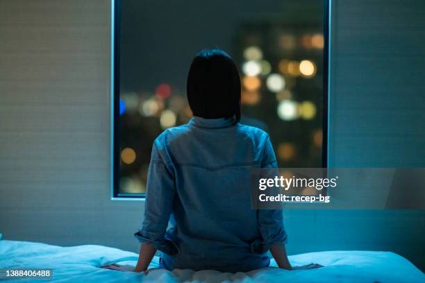 部屋のベッドに一人で座り、夜に窓から覗く女性の背面図 - rear view ストックフォトと画像