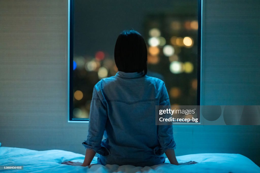 Rear view of woman sitting alone on bed in room and looking through window at night