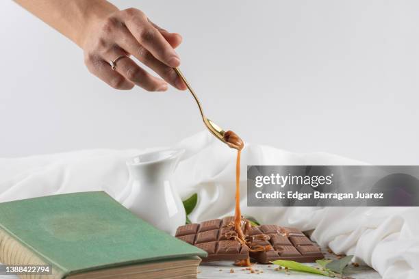 woman's hand holding a spoon while pouring cajeta over a chunky dark chocolate bar on a table with classic decor. - caramel block stock pictures, royalty-free photos & images