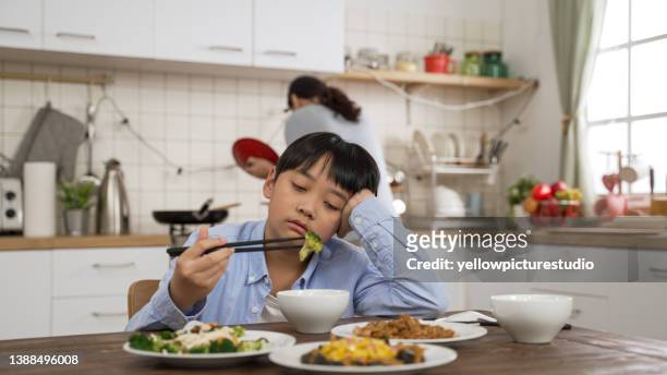 selective focus of upset asian preschool boy holding face with a look of hate as he picks up vegetable from his bowl at dining table and his mother is cooking in kitchen at background - angry parent mealtime stock pictures, royalty-free photos & images