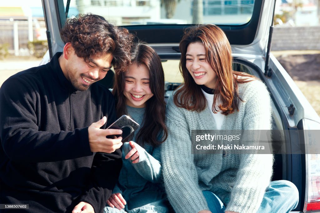 Three friends sitting behind the car and looking at their smartphones.