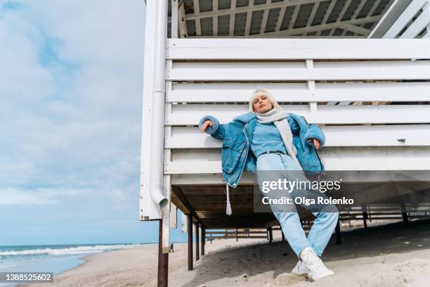 young blond short-haired woman in jeans clothes and a scarf standing and posing near white wooden changing cubicle at the sandy sea beach - denim jacket mockup stock pictures, royalty-free photos & images