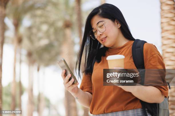 smiling young woman with travel mug standing in palm tree alley with backpack and using mobile app on city break - beautiful filipina stock pictures, royalty-free photos & images