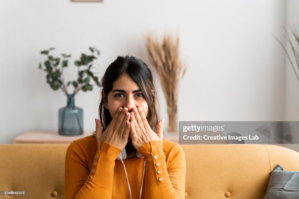 Hispanic woman at home, with a surprised expression covering her mouth with her hands for making a mistake.