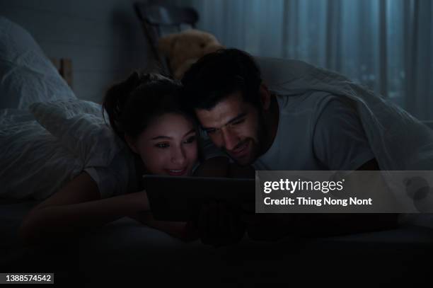 young couple play with tablet or phone on bed in before bedtime at night. relaxed man and smiling woman enjoying with benefits of technology. - young couple at movie together imagens e fotografias de stock