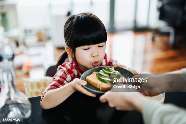 lovely little asian girl sitting at the table in kitchen, making funny faces while being served a healthy breakfast by her mother, avocado with egg on toast in the morning - mutter kind brot glücklich stock-fotos und bilder