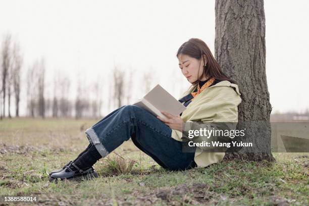 a young asian woman sitting on the grass outdoors and reading a book with her back against a tree - mujer leyendo libro en el parque fotografías e imágenes de stock