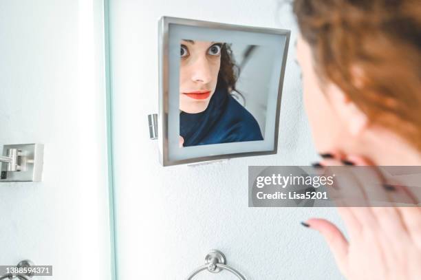 woman in a domestic home bathroom looks into a vanity mirror with a scowl, unhappy with her appearance maybe - body dysmorphia stock pictures, royalty-free photos & images