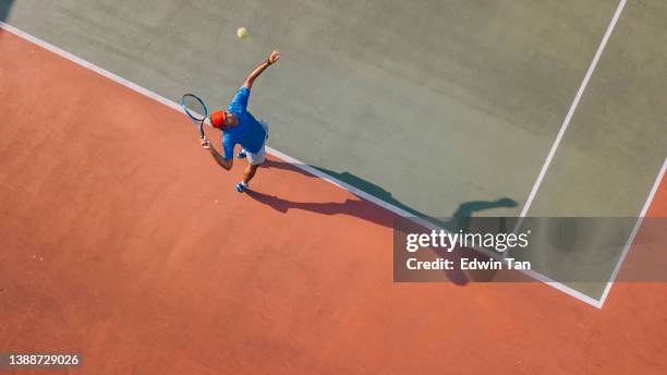 drohnen-blickwinkel asiatischer tennisspieler serviert den ball mit schatten direkt darüber - tennis court top view stock-fotos und bilder