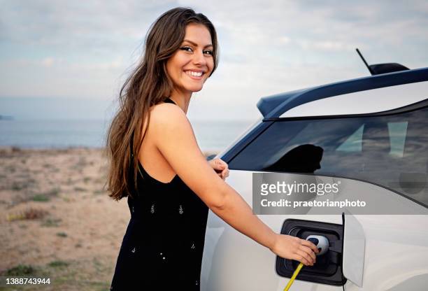 retrato de una joven cargando su coche eléctrico en la playa - red car wire fotografías e imágenes de stock