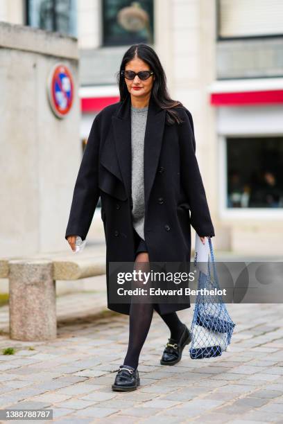 Guest wears black sunglasses, silver earrings, a gray oversized wool pullover, a black oversized long coat, black tights, a black skirt, purple...