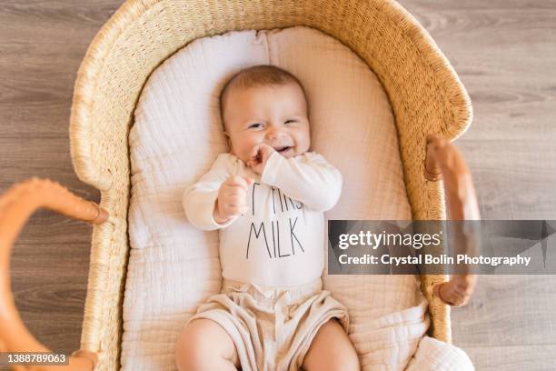 a happy 14 week old baby boy wearing a white onesie with the words 'but first milk' laying in a cozy cream cotton blanket in a seagrass moses basket - laughing teen stockfoto's en -beelden