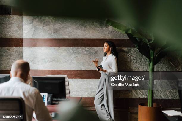 confident female financial advisor talking through in-ear headphones leaning on wall at office - in ear headphones stock pictures, royalty-free photos & images