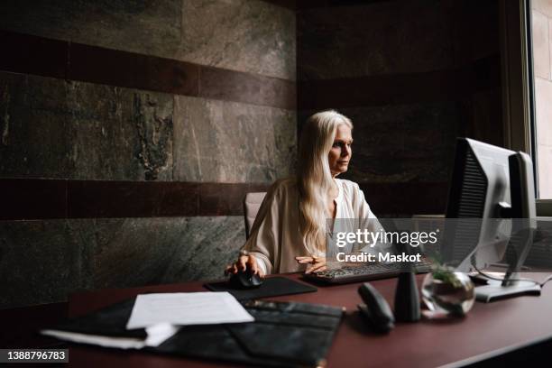 confident mature businesswoman with long white hair using computer at desk in office - ceo desk imagens e fotografias de stock