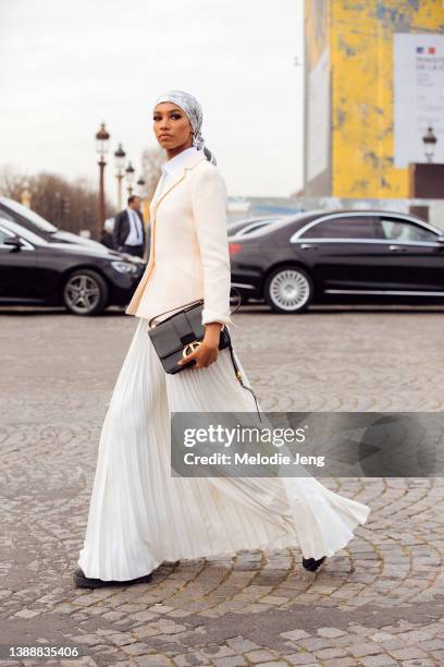 Hijabi model Ikram Abdi Omar wears a white Dior print head scarf, white blazer, black Dior bag, and long cream pleated skirt at the Dior show at...
