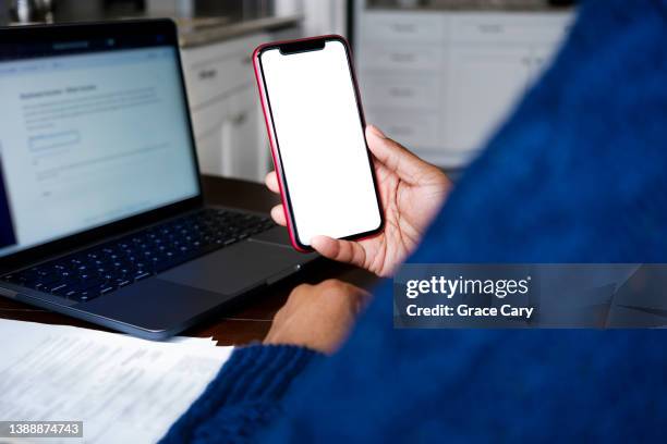 woman holds smart phone with blank screen while working from home on laptop - laptop and iphone mockup stock pictures, royalty-free photos & images