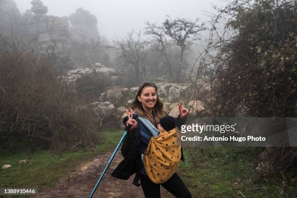 Young woman carrying her baby in a baby carrier while hiking in the mountains. Foggy day.