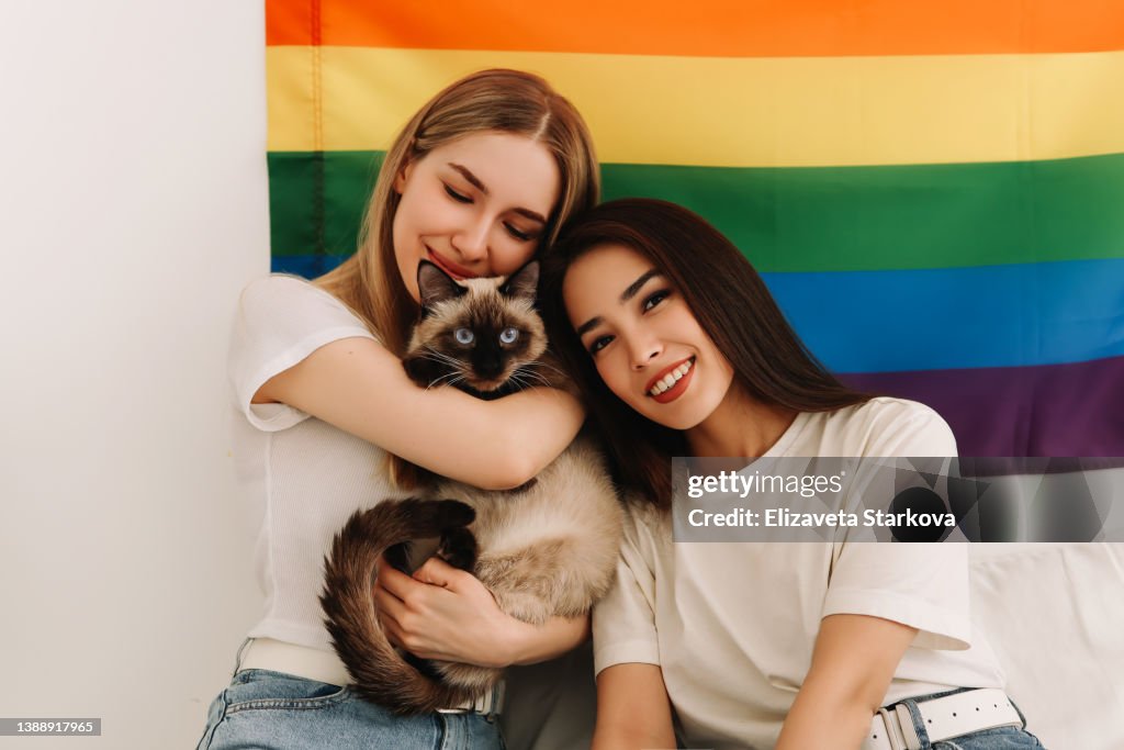 Two happy smiling lesbian gay couple girls in white T-shirts hug and kiss their child's pet cat sitting on white bedding on the bed in the bedroom against the background of a rainbow flag symbol of the LGBT community in the cozy interior of the house.