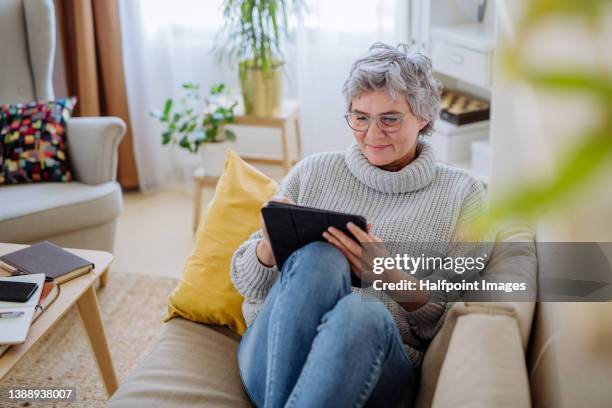 mature woman using digital tablet while sitting on sofa at home - e reader stock pictures, royalty-free photos & images