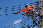 Electrician with safety equipment and work tools is installing cable lines and electrical system on electric power pole against blue sky