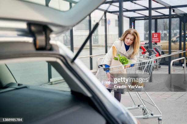 beautiful woman loading groceries into her car in a supermarket parking lot - shopping trolleys stockfoto's en -beelden