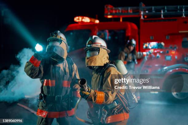 firefighters shining a searchlight while holding a hose next to a fire engine - brandweeruniform stockfoto's en -beelden