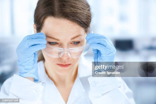 a young female scientist in a white lab coat adjusts her protective glasses - research foundation stock pictures, royalty-free photos & images