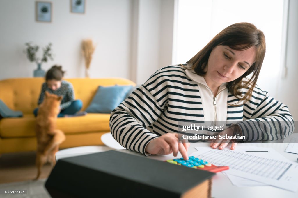 Woman managing the monthly family budget while her daughter plays with a dog in the background.