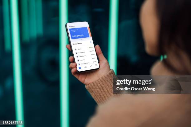 young woman checking credit card transactions on mobile phone against futuristic holographic background - mirar por encima del hombro mujer fotografías e imágenes de stock