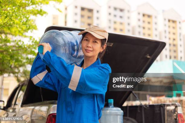 reife wasserlieferfrau in blauer uniform und baseballkappe, die während der lieferung zu hause und im büro einen großen wasserbehälter trägt - gallone stock-fotos und bilder