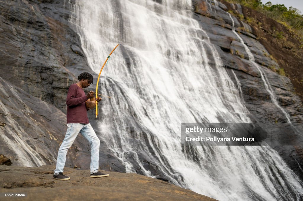 Man playing berimbau beside waterfall