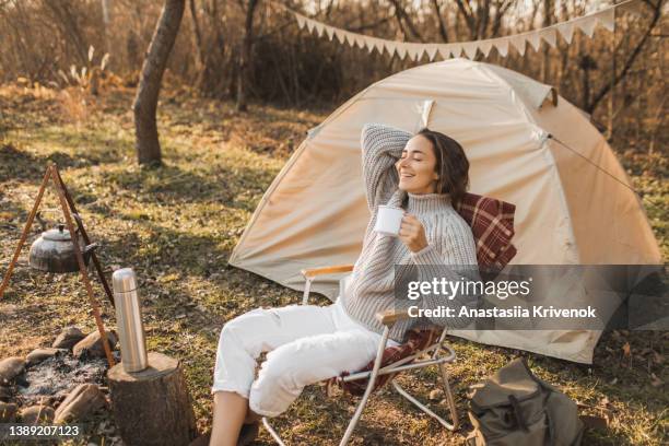 woman drinking tea and sitting in folding chair near camp tent outdoors. - eco tourism stock pictures, royalty-free photos & images