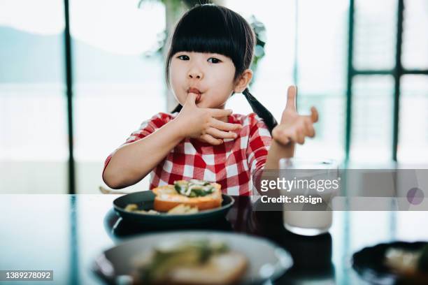 lovely little asian girl licking her finger while having healthy breakfast, avocado with egg on toast in the morning. kids healthy eating habit and lifestyle concept - girls licking girls stock pictures, royalty-free photos & images
