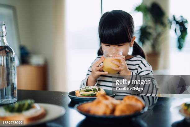 little asian girl sitting at the table in kitchen drinking a glass of fresh orange juice while having breakfast in the morning. kids healthy eating habit and lifestyle concept - orangensaft stock-fotos und bilder