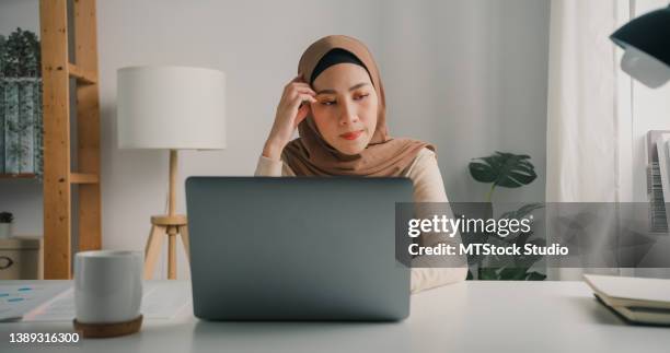 young asian muslim businesswoman wearing hijab working on a laptop sitting at desk in home. - islamic finance stock pictures, royalty-free photos & images
