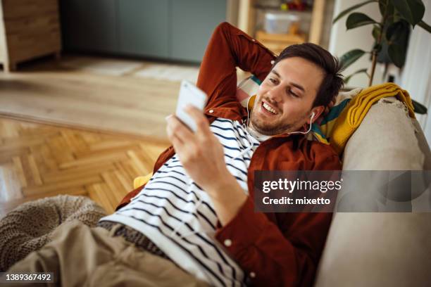 young man listening music and using smart phone on sofa - podcast headphones stock pictures, royalty-free photos & images