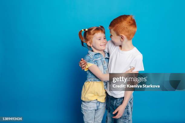 a little boy with red hair hugs a sister girl with red hair on a blue background look at each other and smile - family smile stock-fotos und bilder