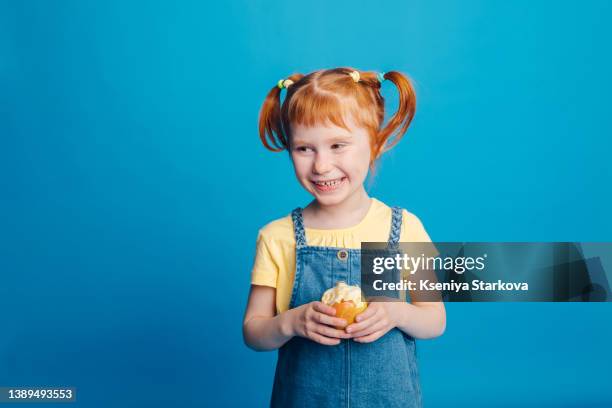 little girl with red hair eats apples on a blue background looks at the camera and smiles - child portrait studio stock-fotos und bilder