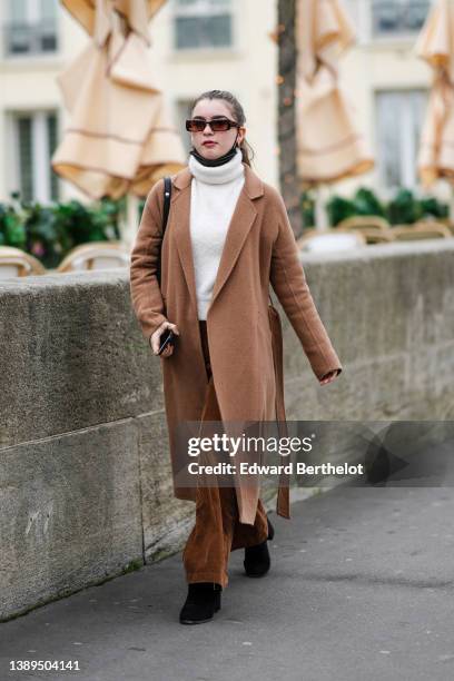 Guest wears sunglasses, a white wool turtleneck pullover, a brown long coat, camel ribbed velvet large pants, black suede block heels ankle boots, a...
